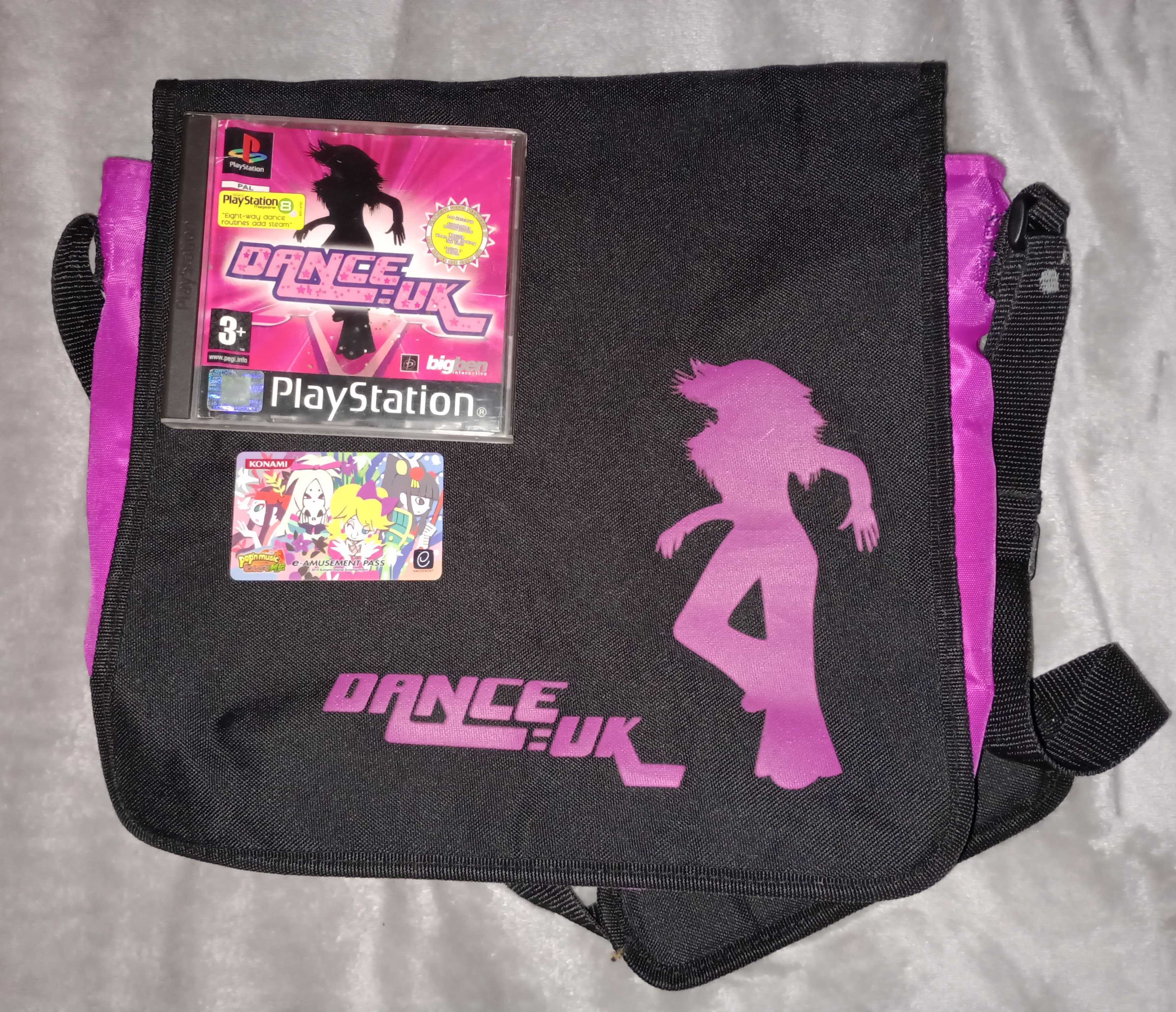 a black shoulder-bag with hot pink details and hot pink text reading 'DANCE:UK' across the front. lying on top of it is the DANCE:UK PS1 game, and TROUT's e-amusement card.
