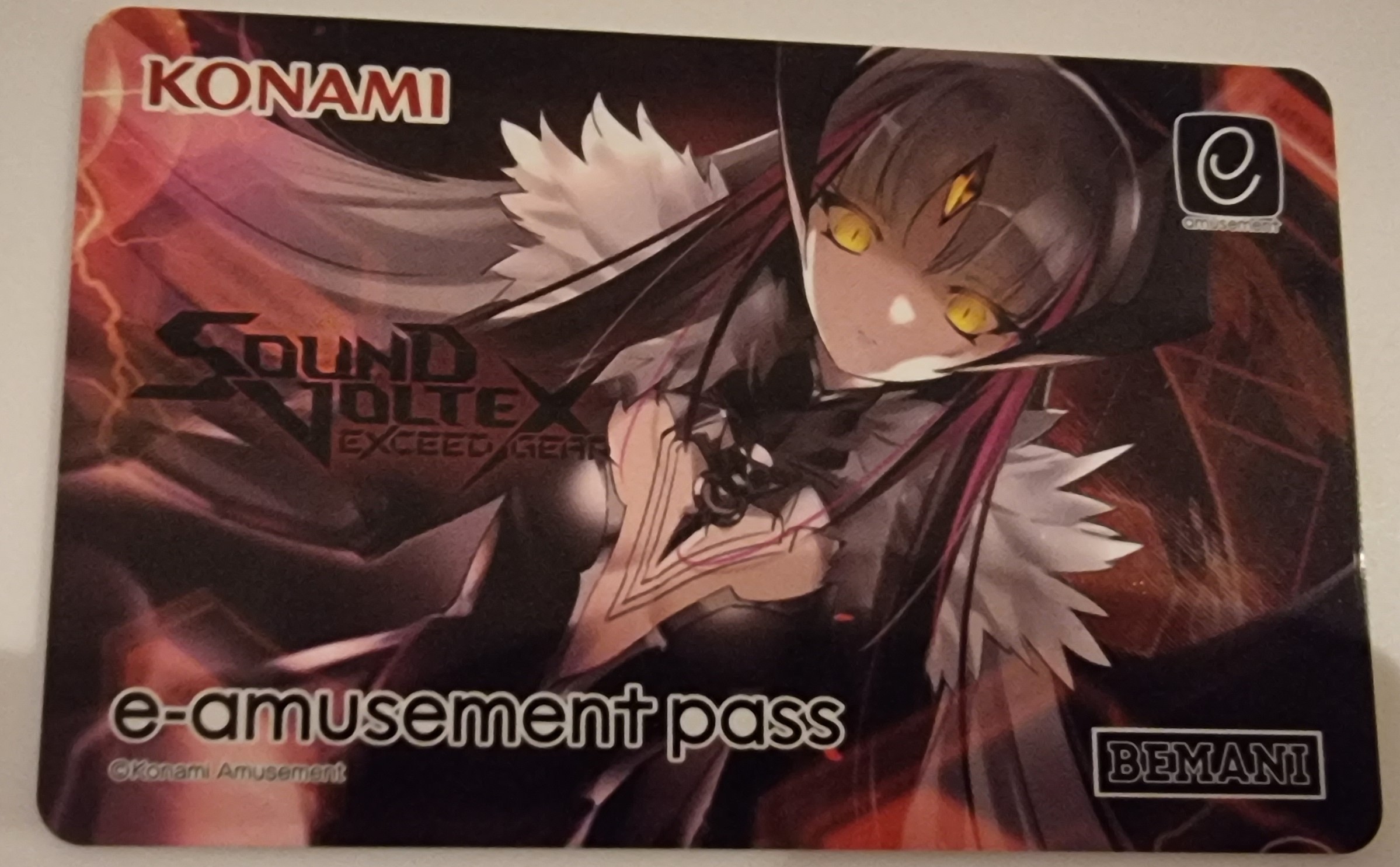 A different e-amusement card. It has a design of a yellow-eyed demon-esque lady on it, with the Sound Voltex game logo to her left.